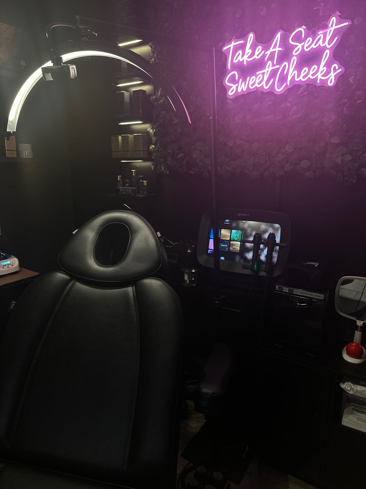 Neon sign reading 'Take A Seat Sweet Cheeks' in a dark room with a chair and mirror at FRZN Aesthetics in Frisco, Texas